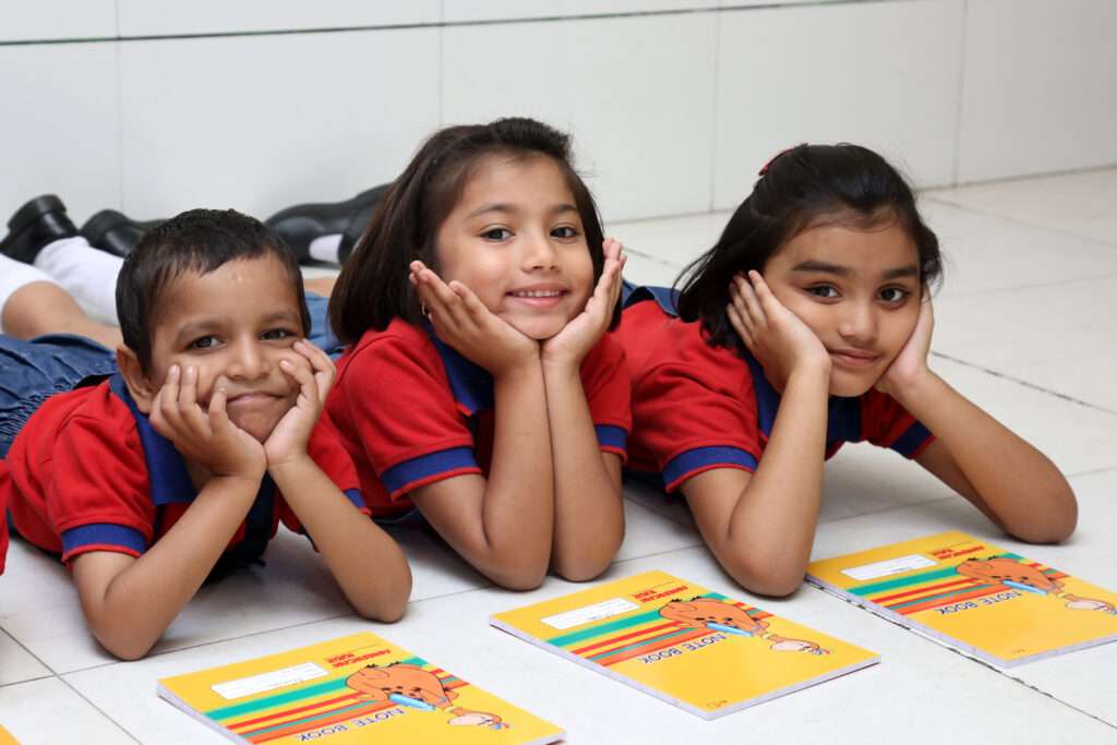 Happy preschool children at American Kidz in Pimple Saudagar, dressed in red uniforms, lying on the floor with notebooks, smiling and enjoying their learning time.
